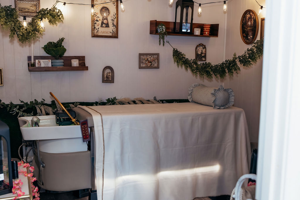 Cozy boho head spa room with vintage art, eucalyptus garland, string lights, draped treatment bed, and gold shampoo basin at A Shade of Vintage in Cheyenne, Wyoming